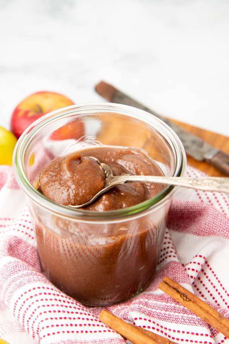 A jar of apple butter with a spoon in it stands on a red and white kitchen towel.