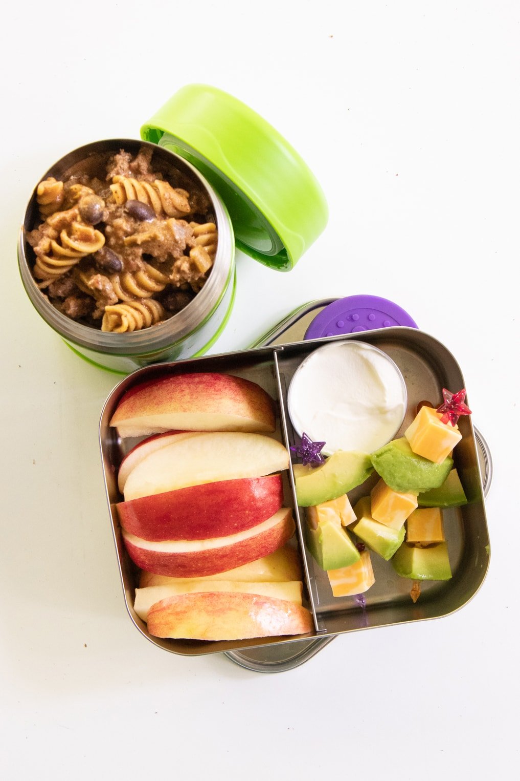 Taco pasta skillet in a thermos next to a bento-style lunch box filled with apple slices, avocado, and cheese.