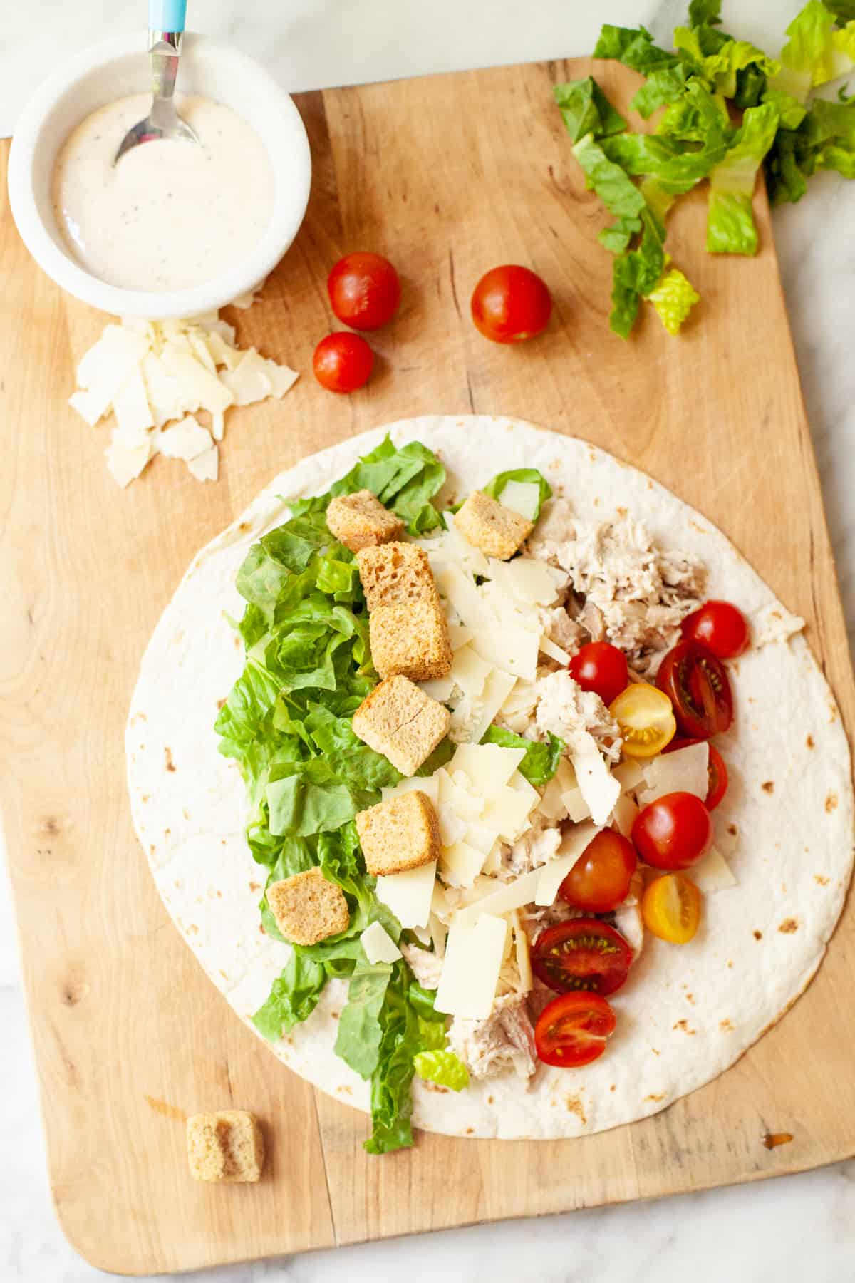 Lettuce, croutons, cheese, chicken, and tomatoes are placed on a tortilla. Dressing and tomatoes also rest on wooden cutting board.