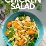 A single serving bowl of mandarin chicken salad sits on a linen napkin surrounded by homemade dressing and garnishes in bowls. A text overlay reads, "Mandarin Chicken Salad."