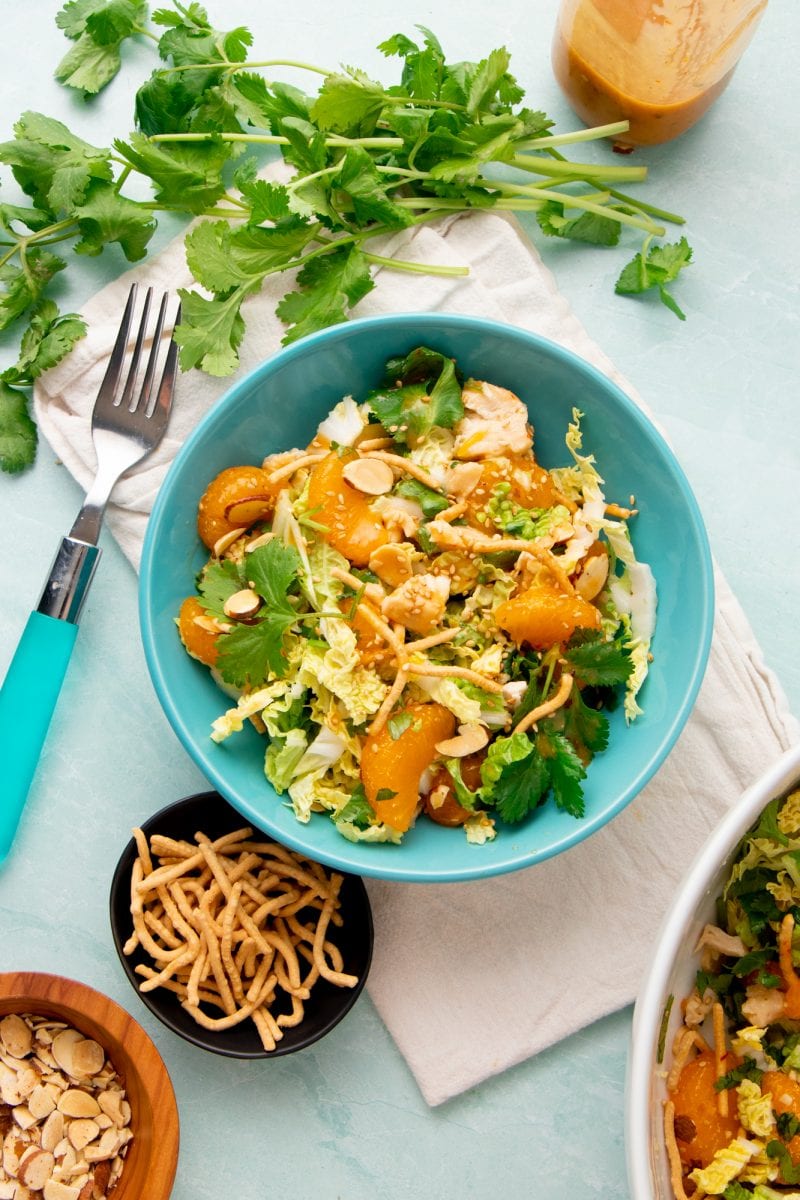 A single serving bowl of mandarin chicken salad sits on a linen napkin surrounded by homemade dressing and garnishes in bowls.