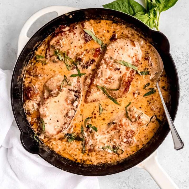 A skillet of marry me chicken garnished with fresh basil rests on a light countertop with a sprig of fresh basil tucked beside it.