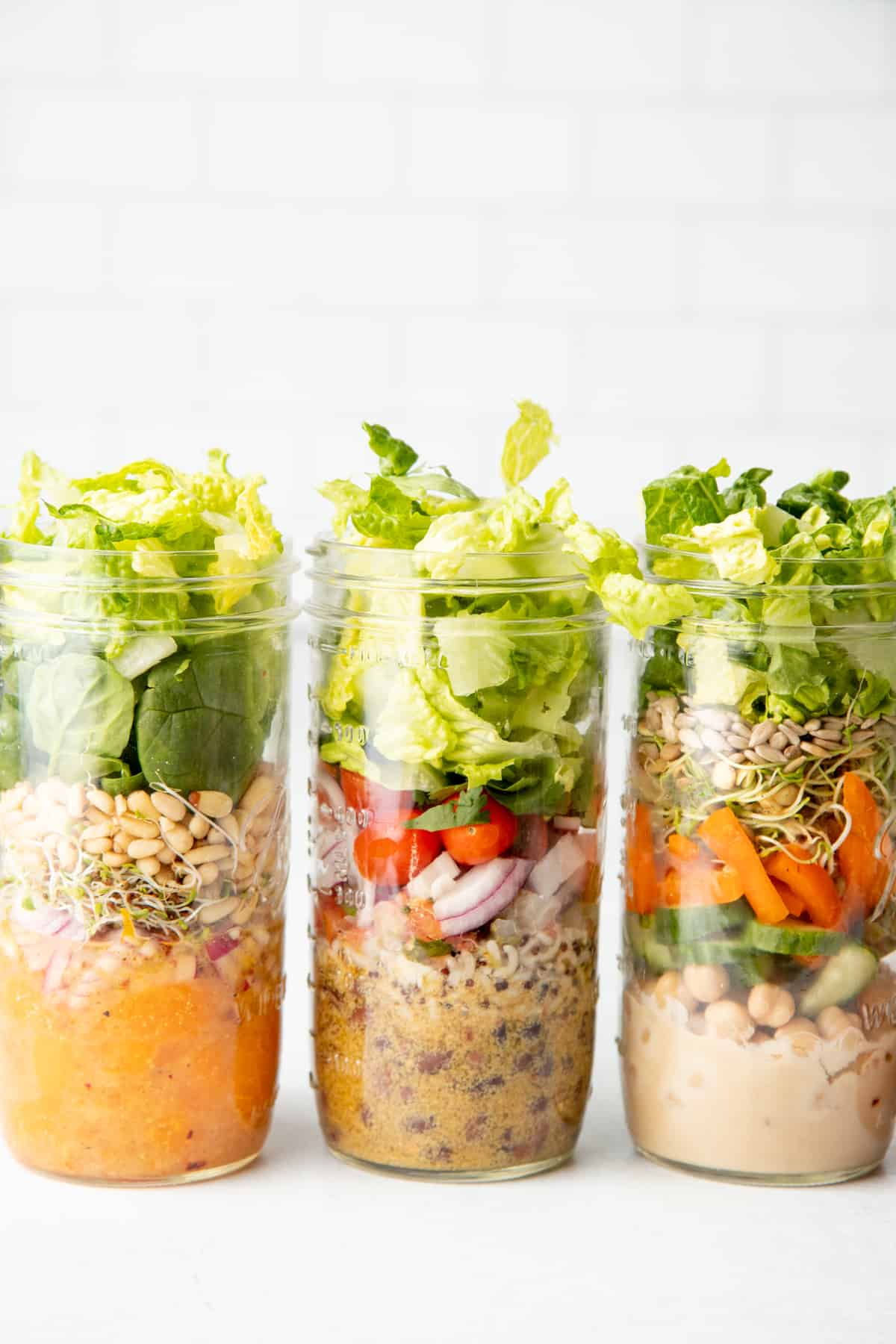 Three tall mason jars sit side-by-side in front of a white wall. Jars are layered with salad ingredients.