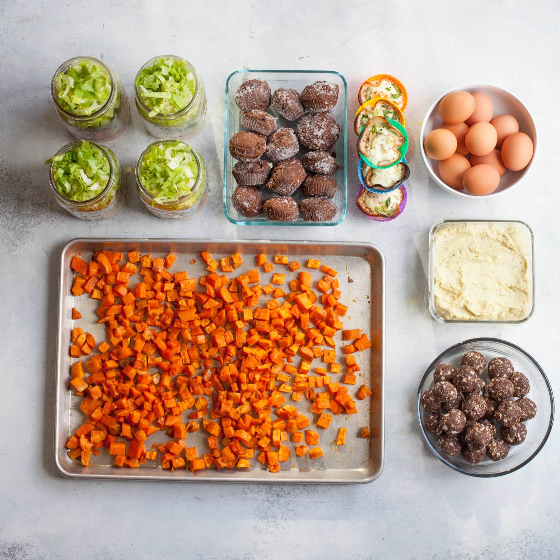 Meal prep components laid out in individual containers - roasted sweet potato cubes, salads in jars, muffins, egg cups, hard boiled eggs, hummus, and date bites.