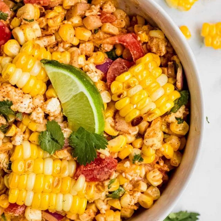 Tight view of a large white serving bowl of creamy corn salad garnished with cilantro and a lime wedge.