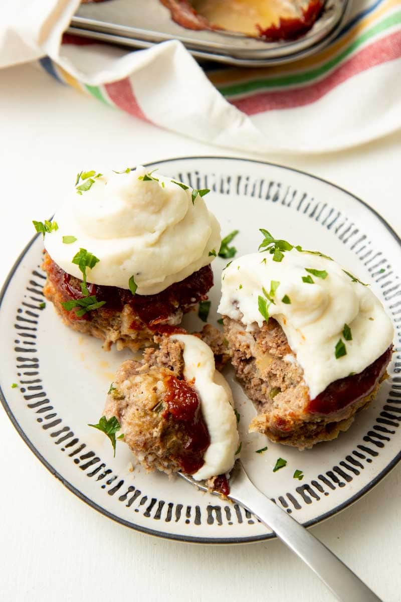 A fork holds a bite of mini meatloaf topped with mashed potatoes on a plate with two mini loaves.