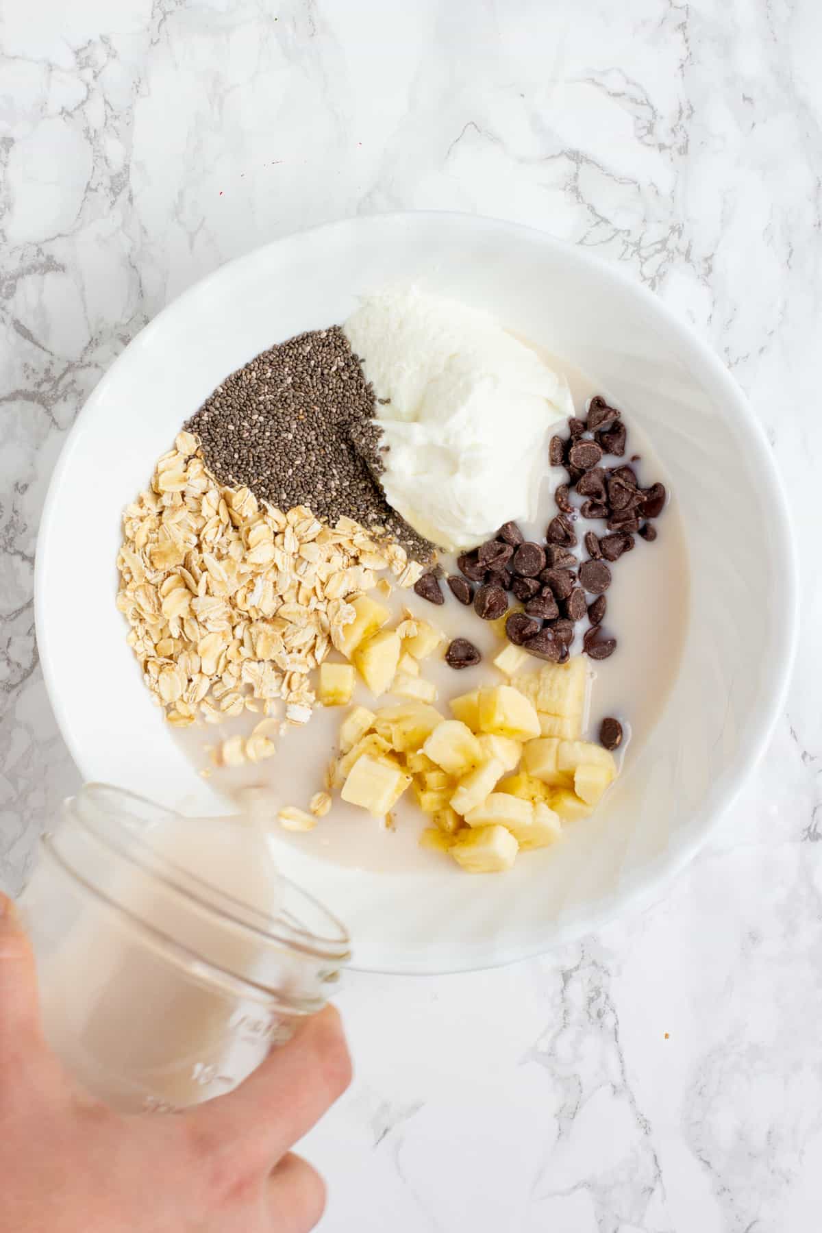 A hand pours milk from a glass jar into a white bowl. The white bowl is filled with other ingredients, including yogurt, chopped banana, chocolate chips, chia seeds, and rolled oats.