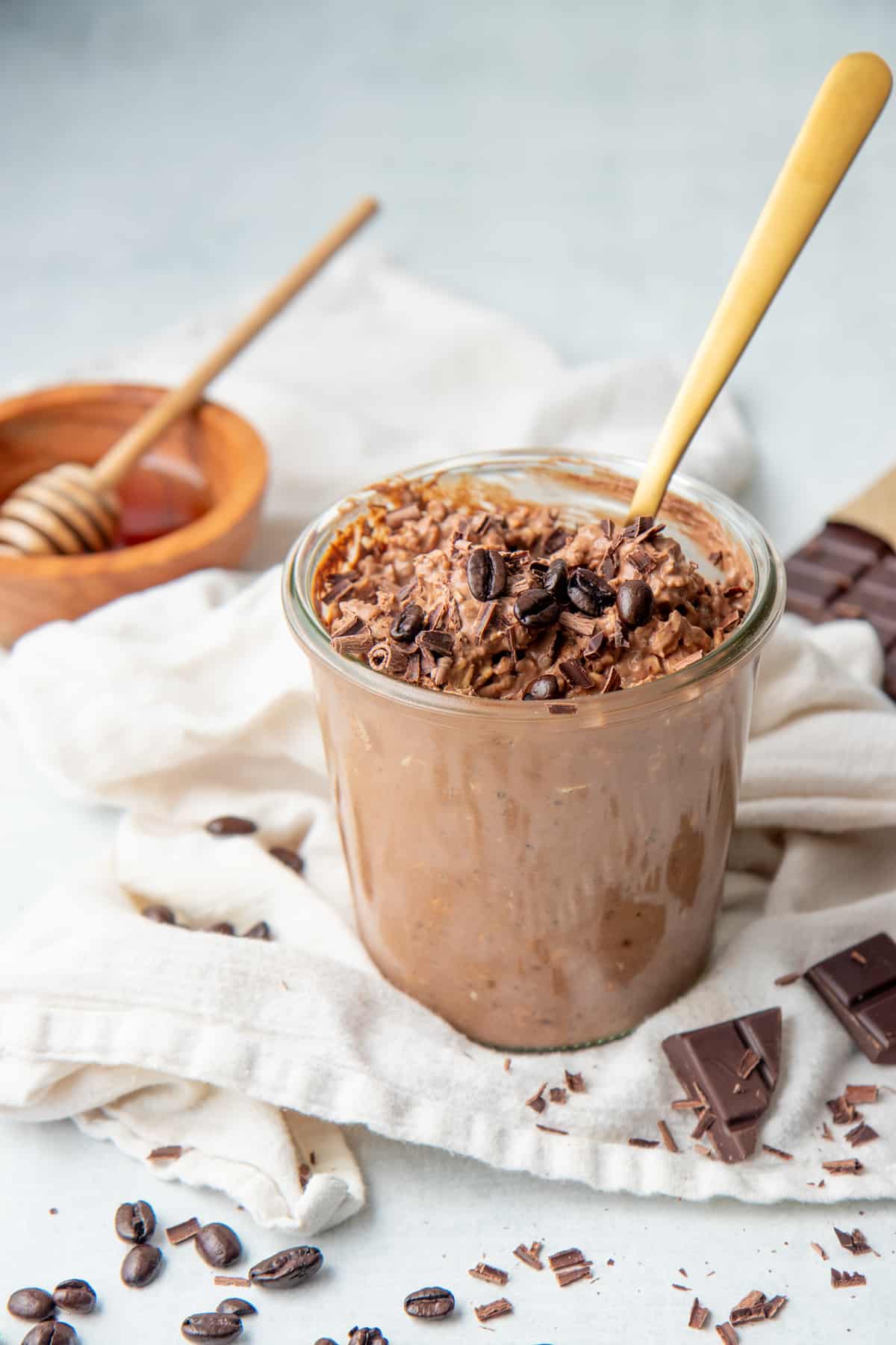 A gold spoon rests in a jar of chocolate coffee overnight oats. A small bowl of honey also sits on the white dishtowel.