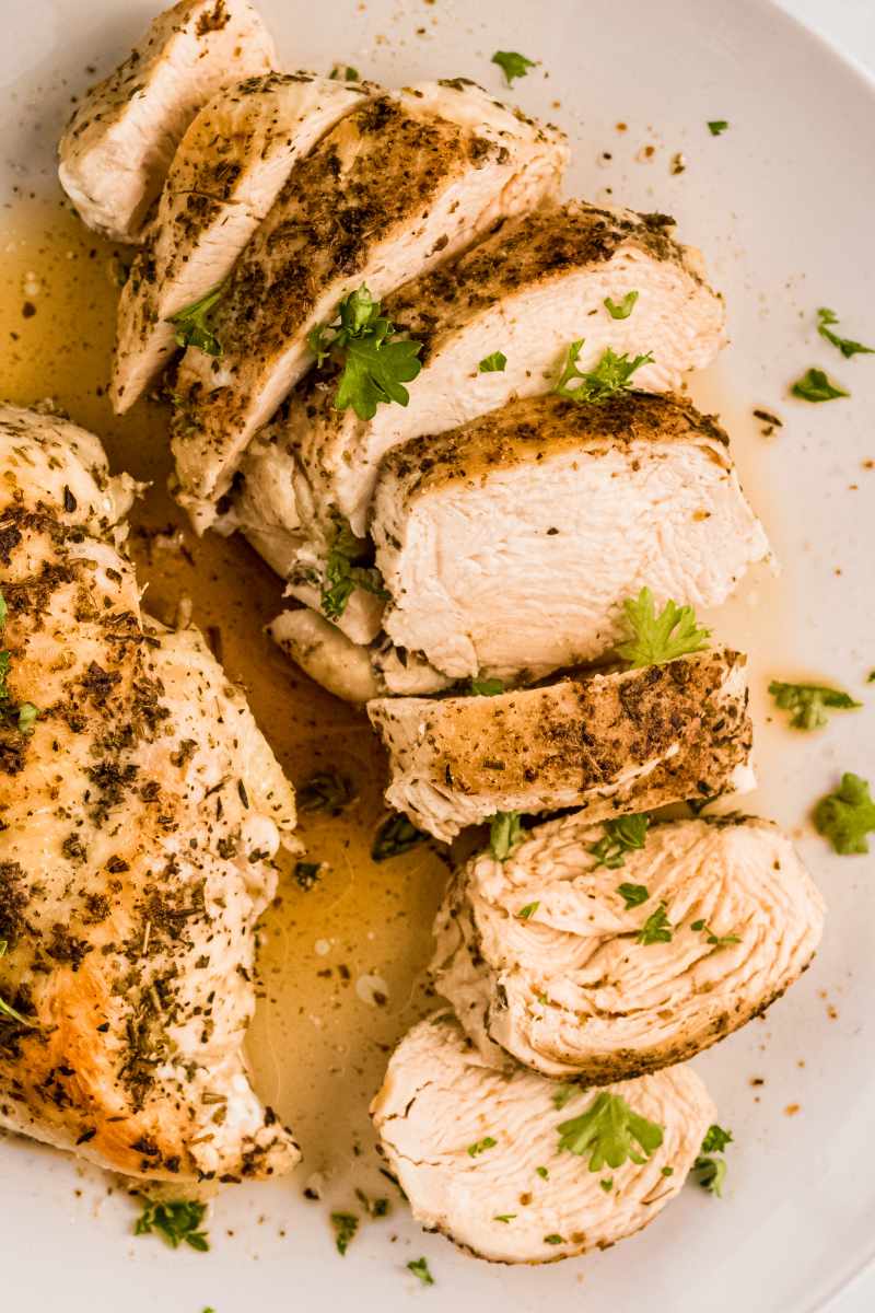 Tight view sliced cooked chicken on a white plate with another chicken breast beside it.