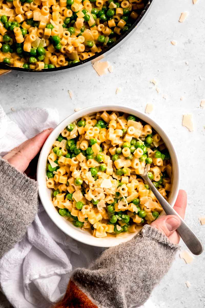 Two hands hug a bowl of pasta with peas served with parmesan and a spoon.