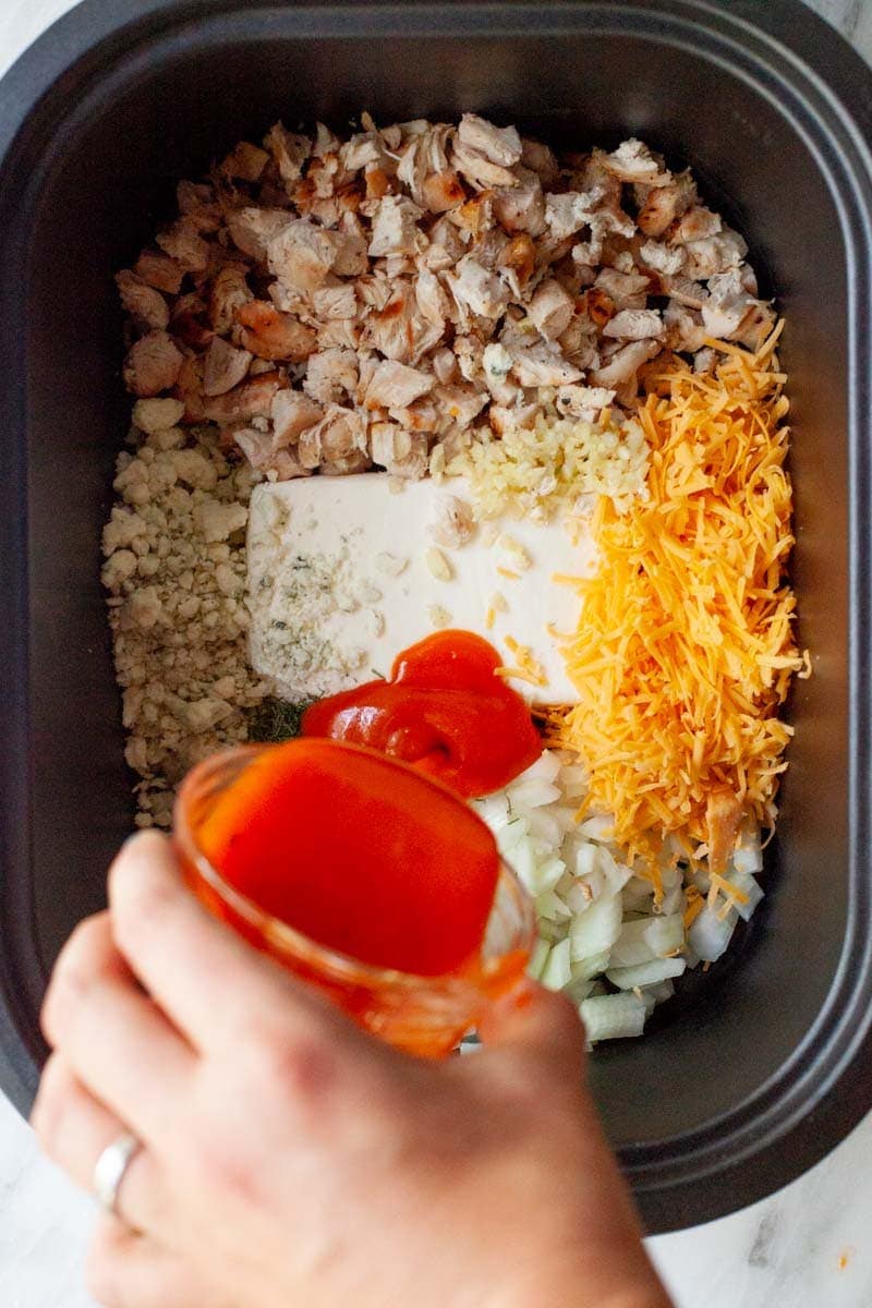 A hand pours buffalo sauce into a slow cooker filled with ingredients including shredded chicken, cream cheese, cheddar cheese, and blue cheese.