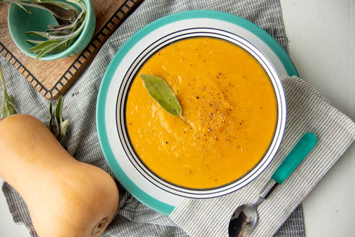 A serving of roasted squash soup garnished with a sage leaf in a bowl on a platter with a spoon and cloth napkin alongside.