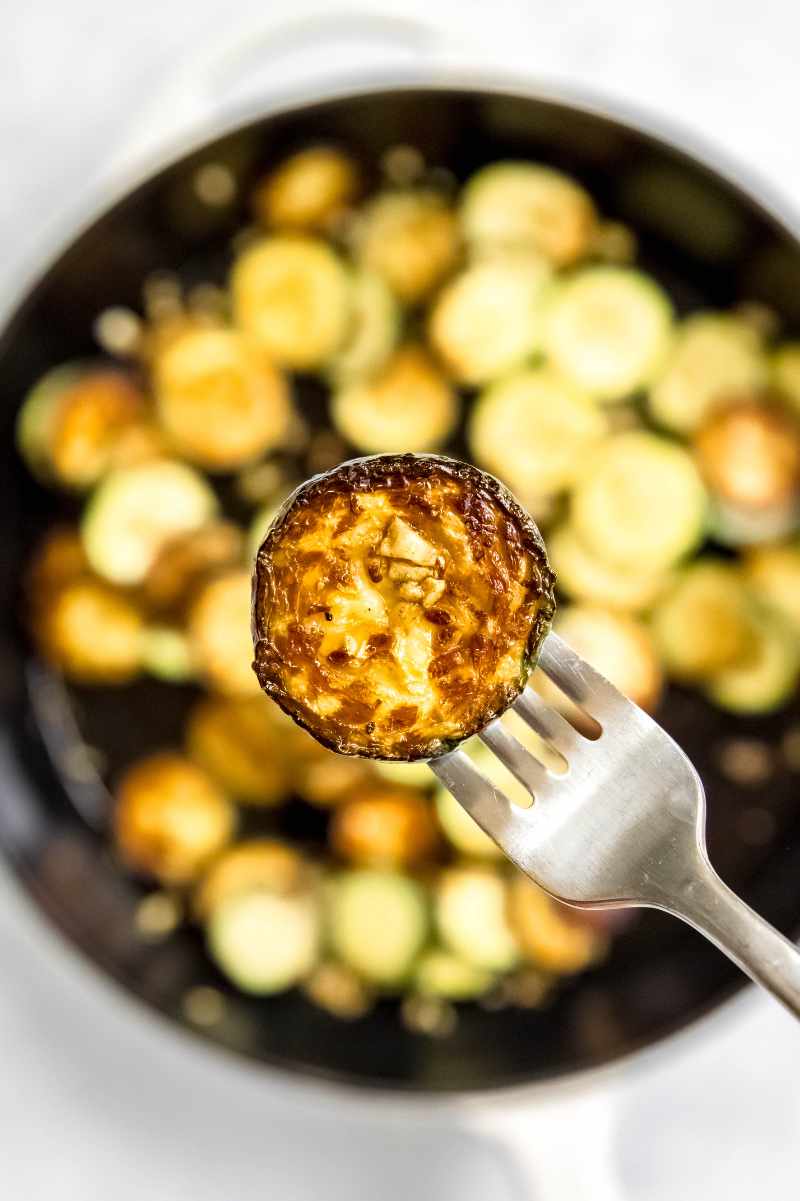 A fork holds a slice of cooked summer squash with garlic above the pan with the rest of the cooked veggies.