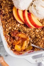 Overhead of a spoon dipping into a peach crisp. Fresh peach slices are arranged on the top of the middle of the crisp.