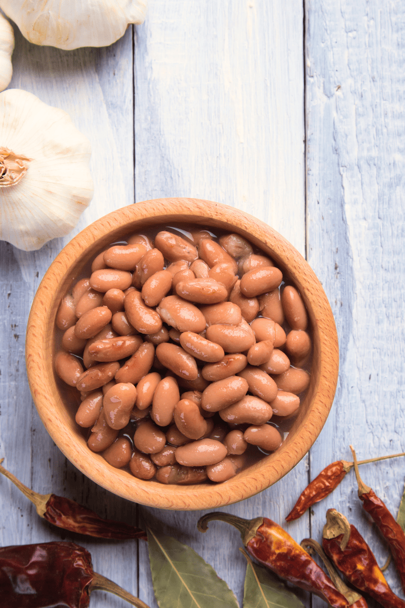 A bowl of pinto beans rests on a grey wooden table surrounded by dried chilies, dried bay leaves, and fresh heads of garlic.