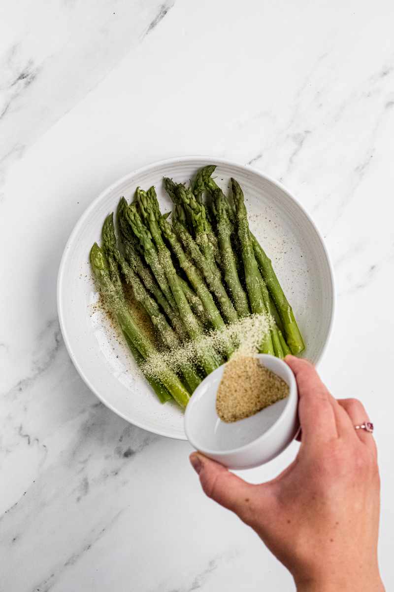 A hand sprinkles dried seasonings from a small white ramekin over perfectly steamed asparagus spears.