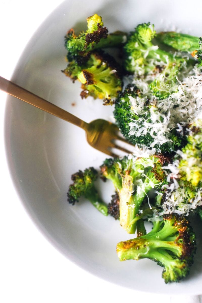 A fork digs into a shallow bowl of broccoli topped with shredded parmesan cheese.