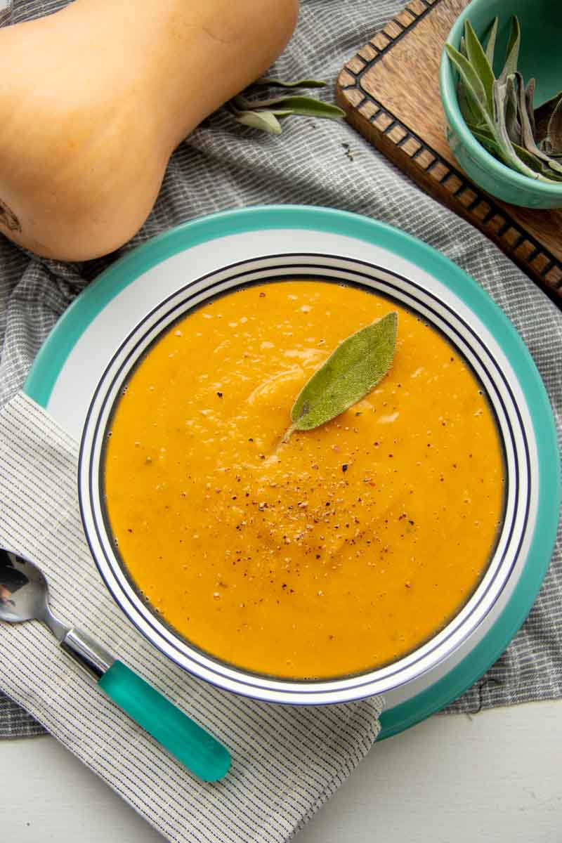 Overhead of a bowl of butternut squash soup served on grey and white fabric with fresh sage and a whole butternut squash nearby.