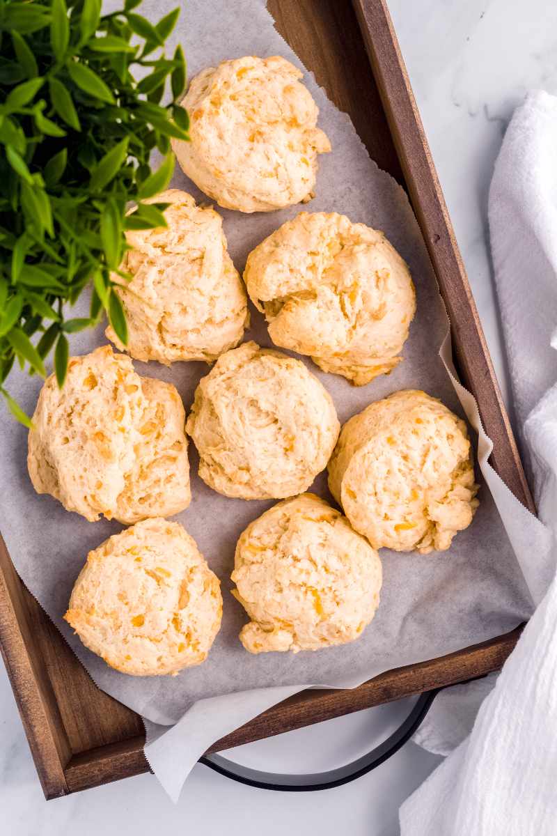 Tender, cheesy drop biscuits sit on a parchment paper lined wooden serving tray.