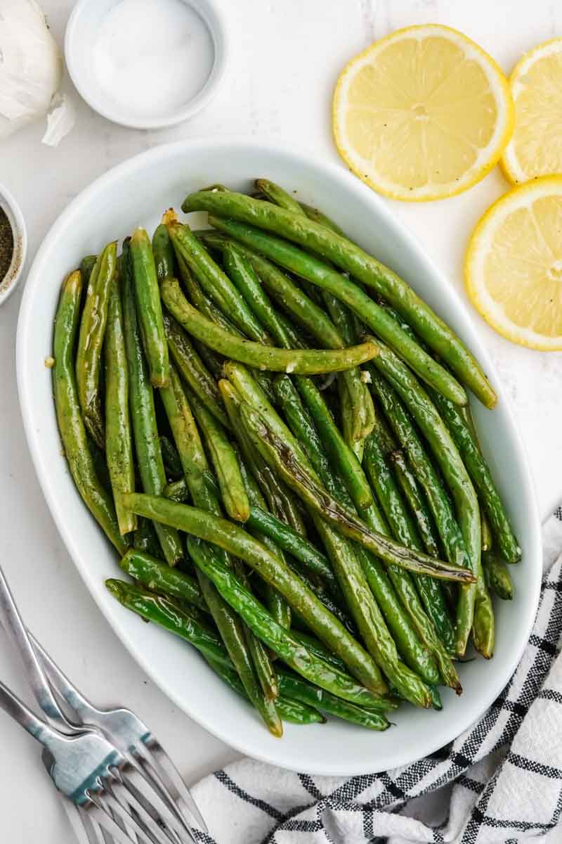 A serving dish of garlic green beans with forks and lemon slices on either side.