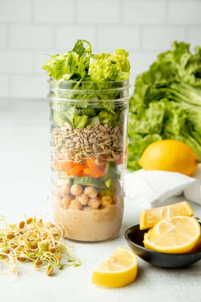 A tall mason jar with layered salad ingredients sits on a white counter in front of a white wall. Lemon slices sit next to the jar in a small black bowl.