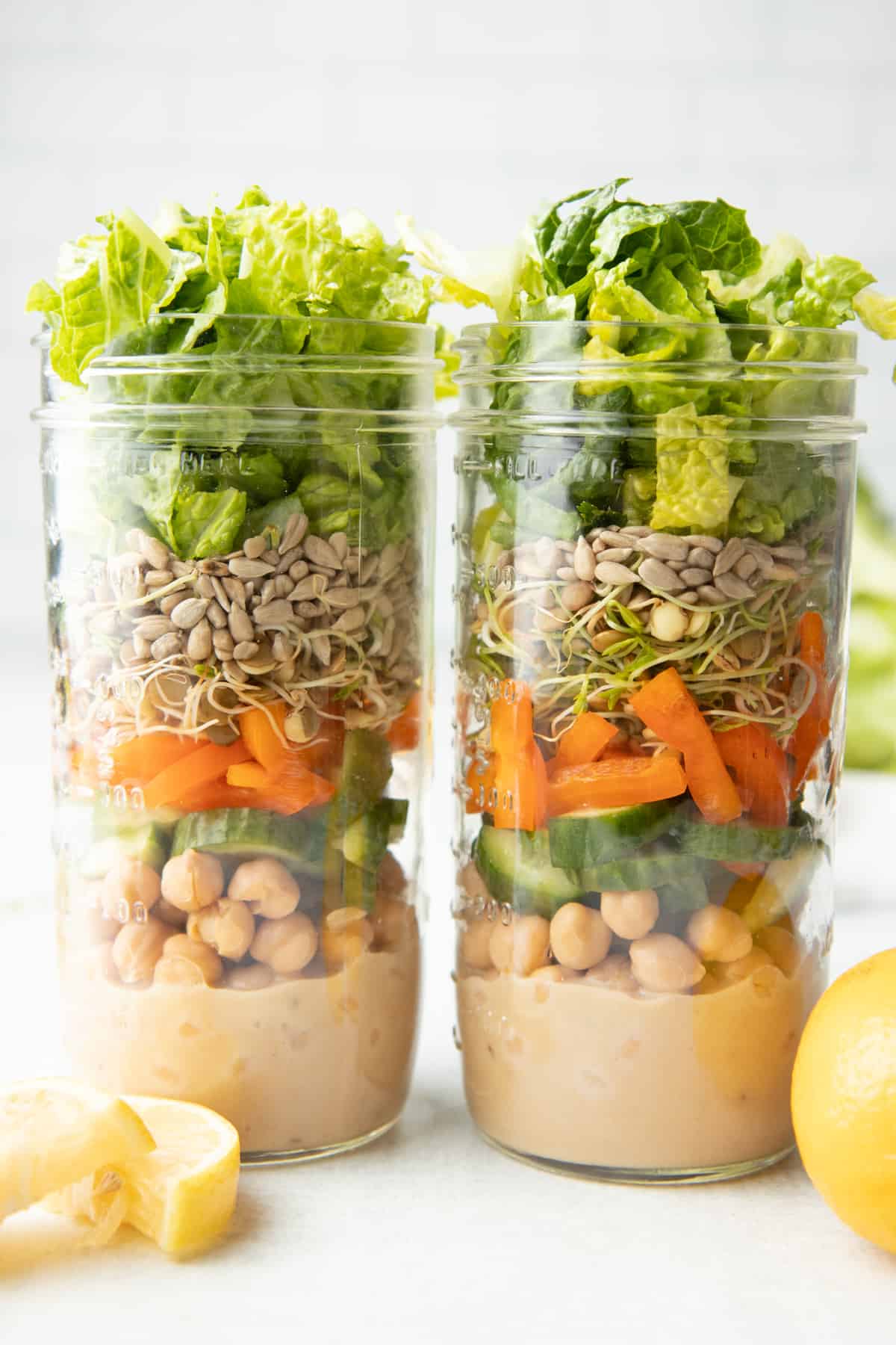 Two tall mason jars sit side by side. Jars are layered with dressing, chickpeas, cut vegetables, sprouts, seeds, and lettuce.