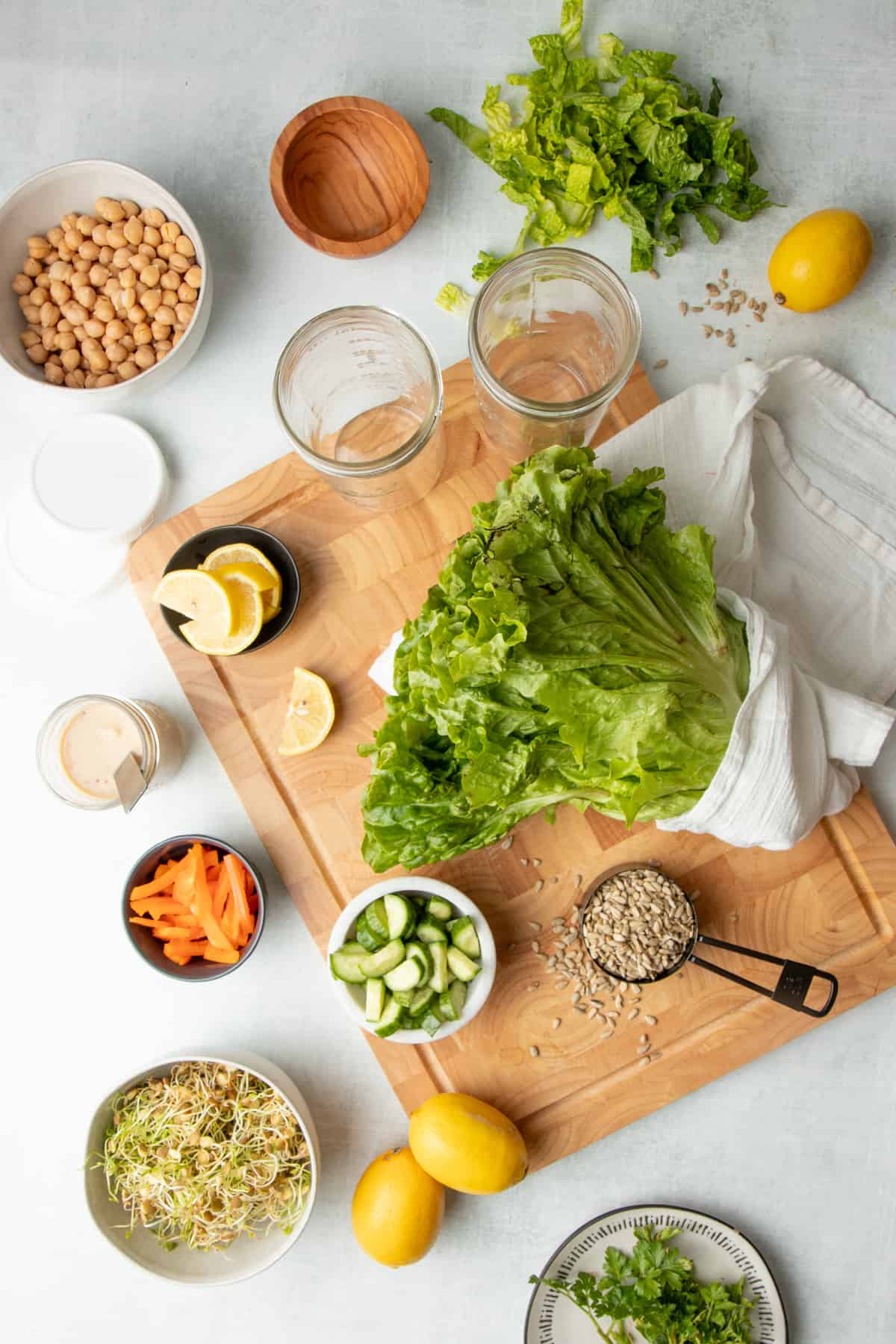 Two empty glass jars, lettuce, and sliced lemons sit on a wooden cutting board. Around the board, chickpeas, sprouts, and other ingredients sit on a white countertop.