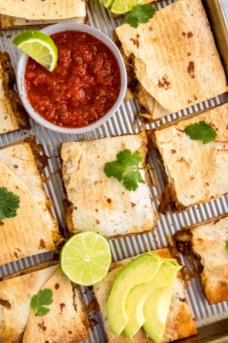 A sheet pan quesadilla dinner made with ground beef is served with salsa, avocado, lime, and cilantro.