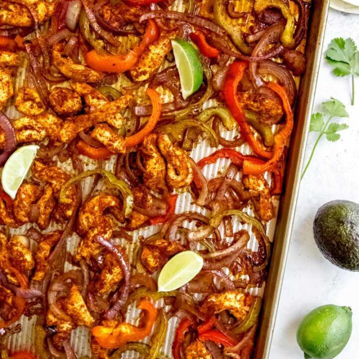 Lime wedges garnish sheet pan chicken fajitas on a counter alongside sprigs of cilantro, flour tortillas, and an avocado.