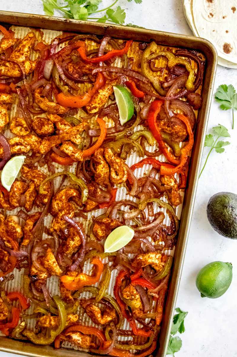 Lime wedges garnish sheet pan chicken fajitas on a counter alongside sprigs of cilantro, flour tortillas, and an avocado.