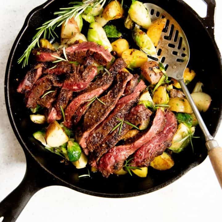 Overhead shot of potatoes and Brussels sprouts in a cast iron skillet, topped with a sliced seared steak