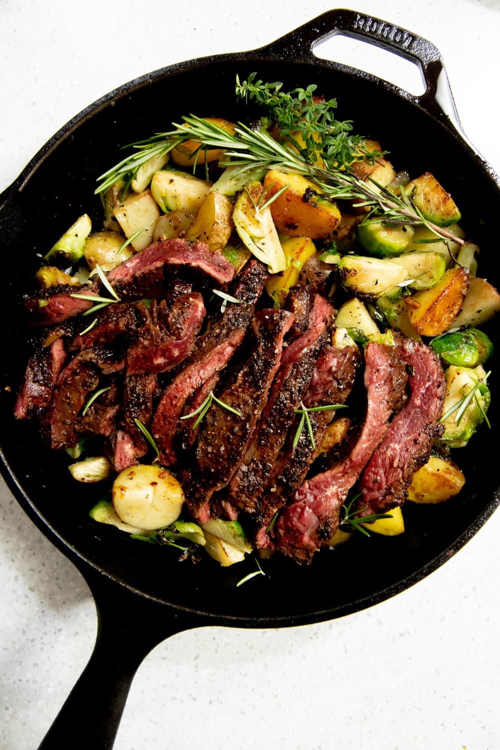 Potatoes and Brussels sprouts in a cast iron skillet, topped with a sliced seared steak