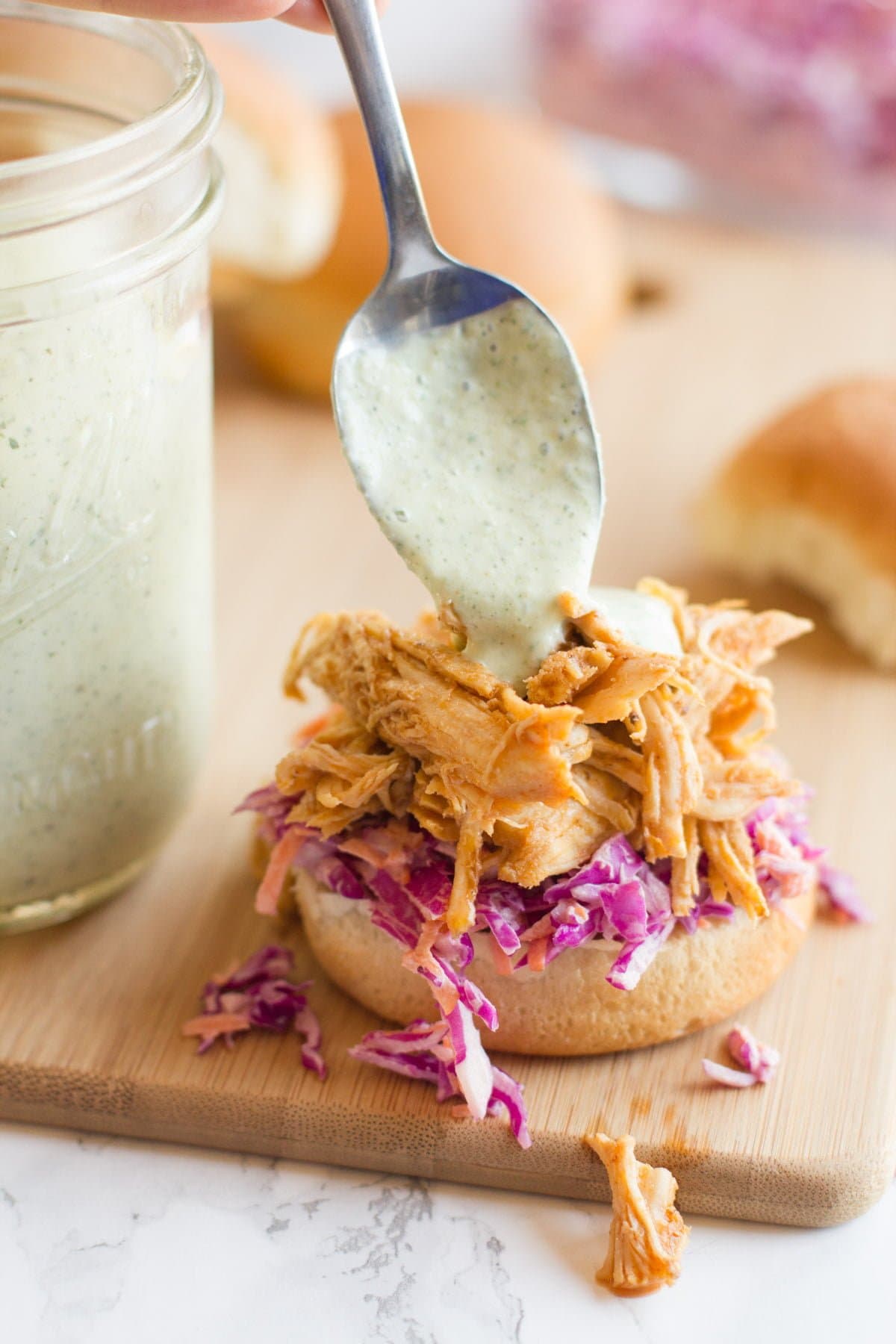 A spoon drizzles greek yogurt ranch sauce onto the top of a sriracha chicken slider.