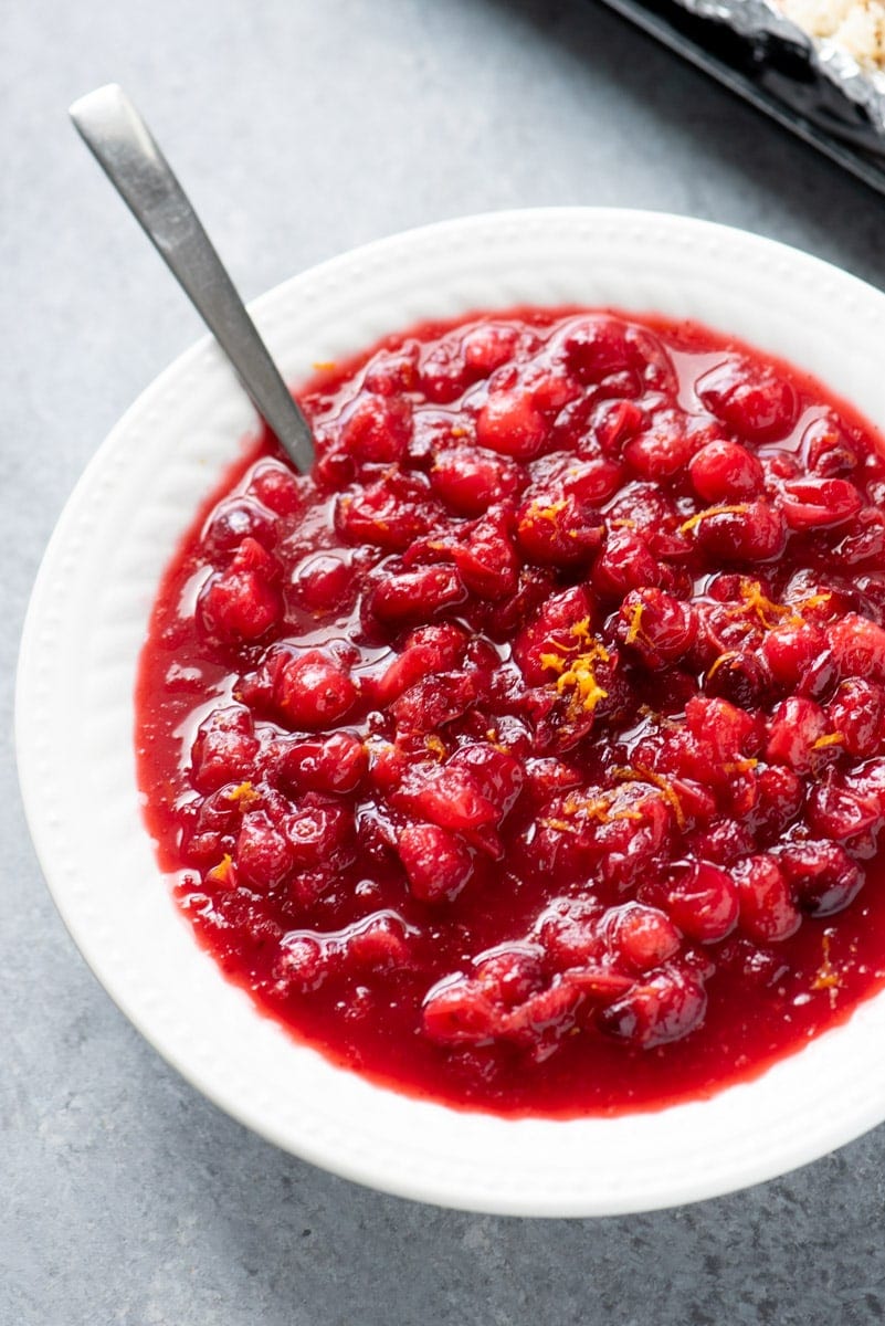 A spoon rests in a white bowl filled with homemade cranberry sauce garnished with citrus zest.