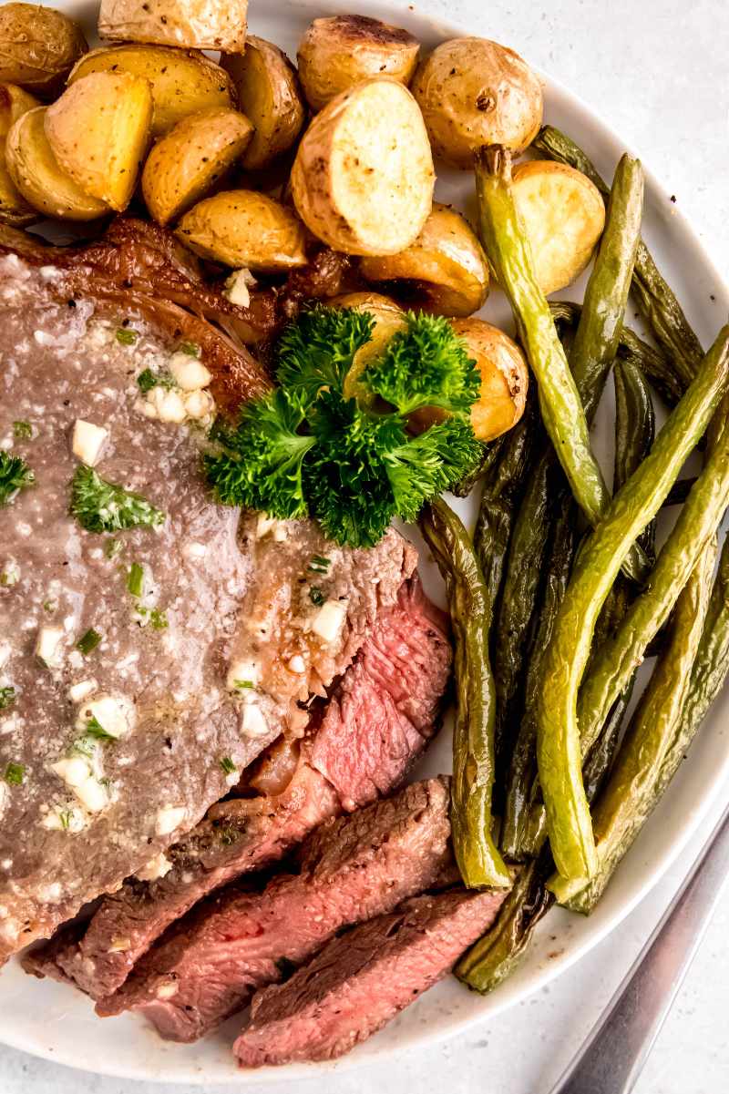 Overhead of a partially sliced garlic butter steak, roasted green beans, and potatoes on a white plate.