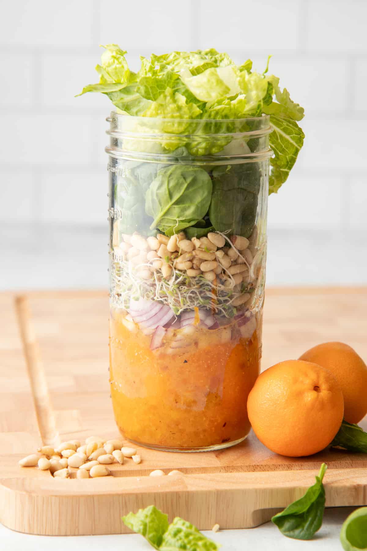 Components for a mandarin salad layered in a mason jar - dressing, mandarin slices, onions, sprouts, pine nuts, and greens. Additional ingredients surround the jar.