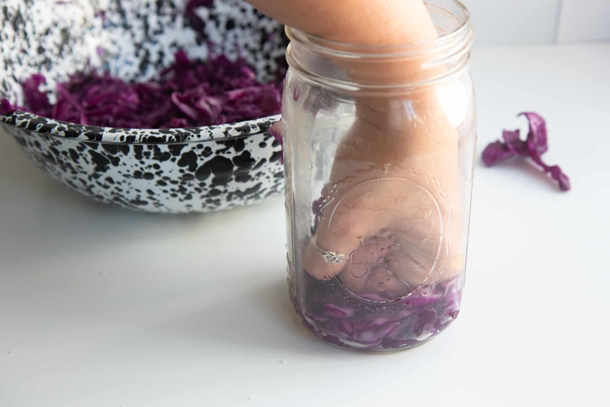 A hand tamps down a fistful of sliced red cabbage in a wide mouth glass mason jar.