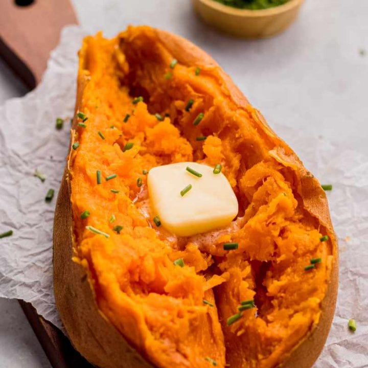 A tender baked sweet potato split down the center with a pat of butter and chopped chives sits on a parchment lined serving board.