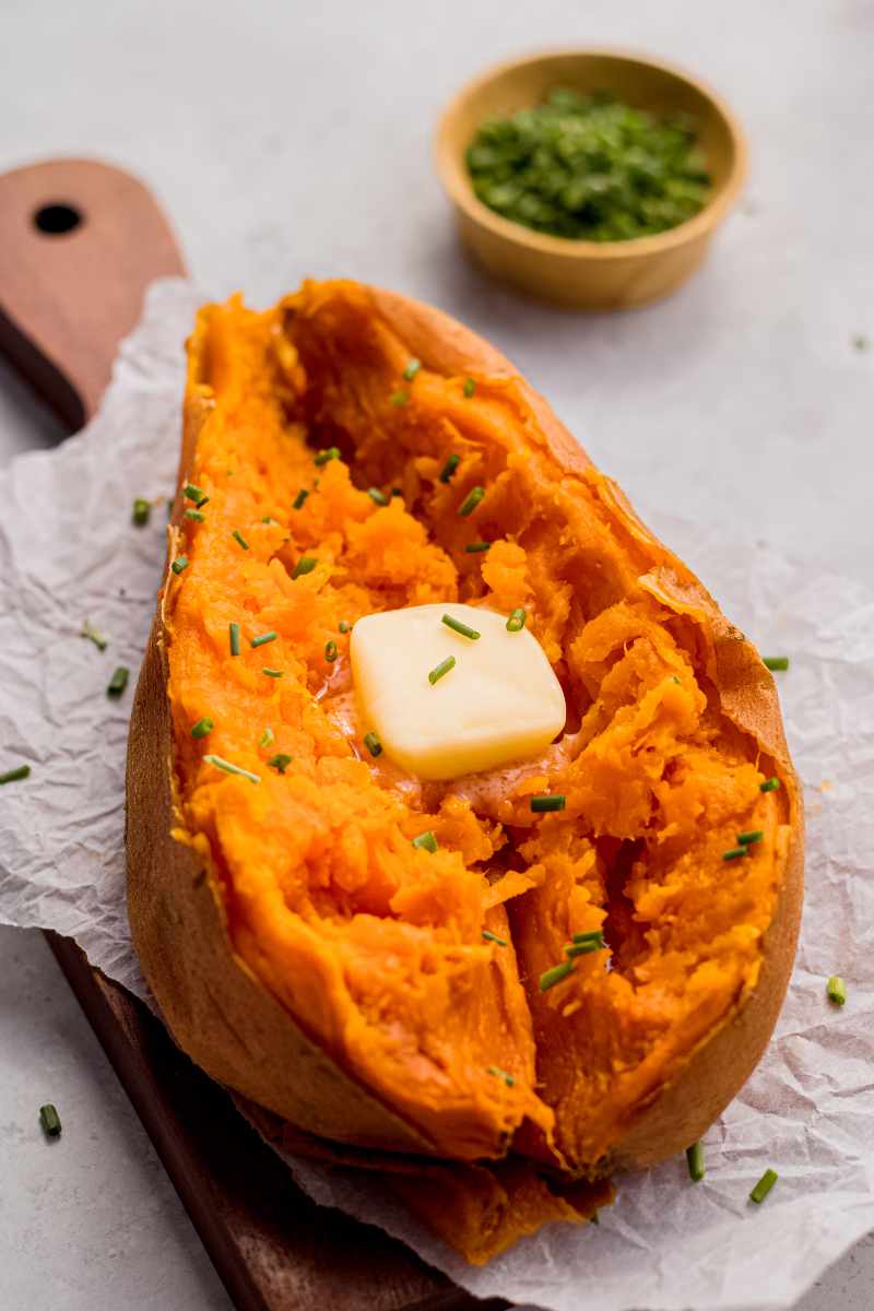 A tender baked sweet potato split down the center with a pat of butter and chopped chives sits on a parchment lined serving board.