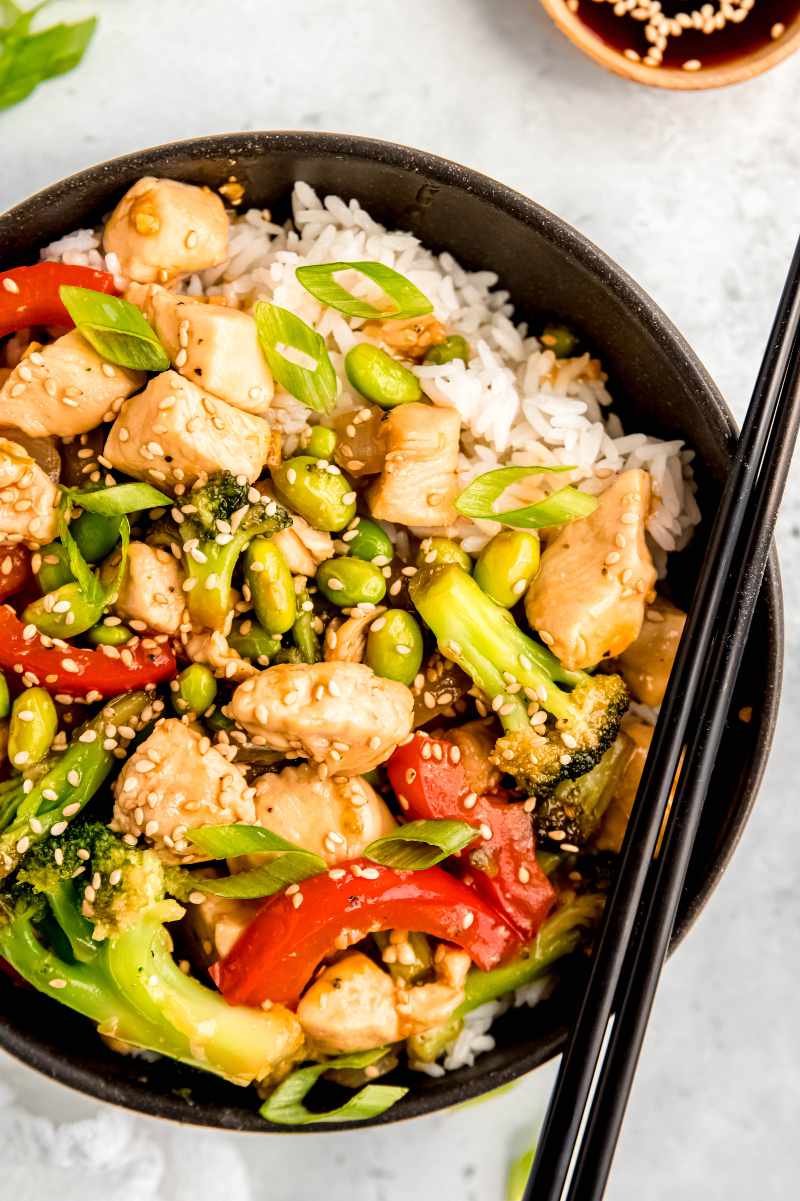 Close view of chopsticks resting along the side of a dark bowl filled with white rice topped with teriyaki chicken and vegetables.