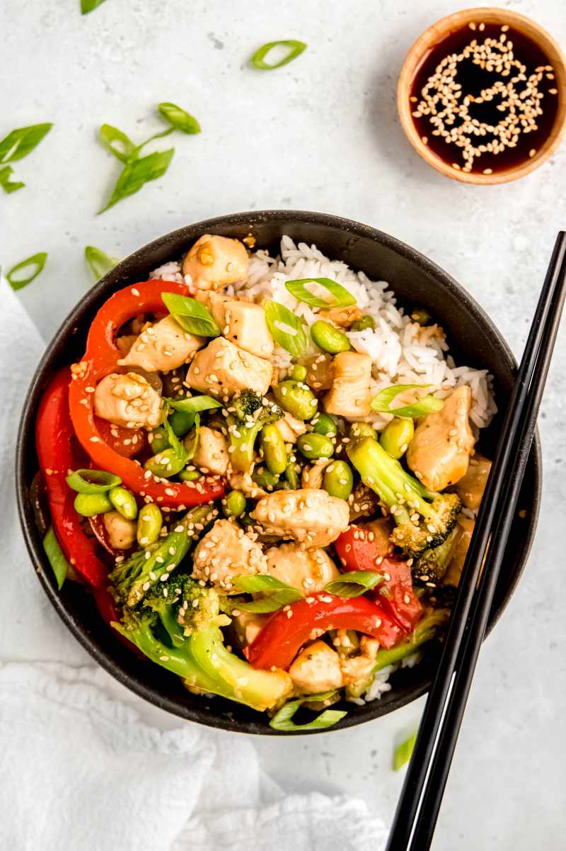 A teriyaki chicken rice bowl served with chopsticks and a small bowl of extra sauce alongside.