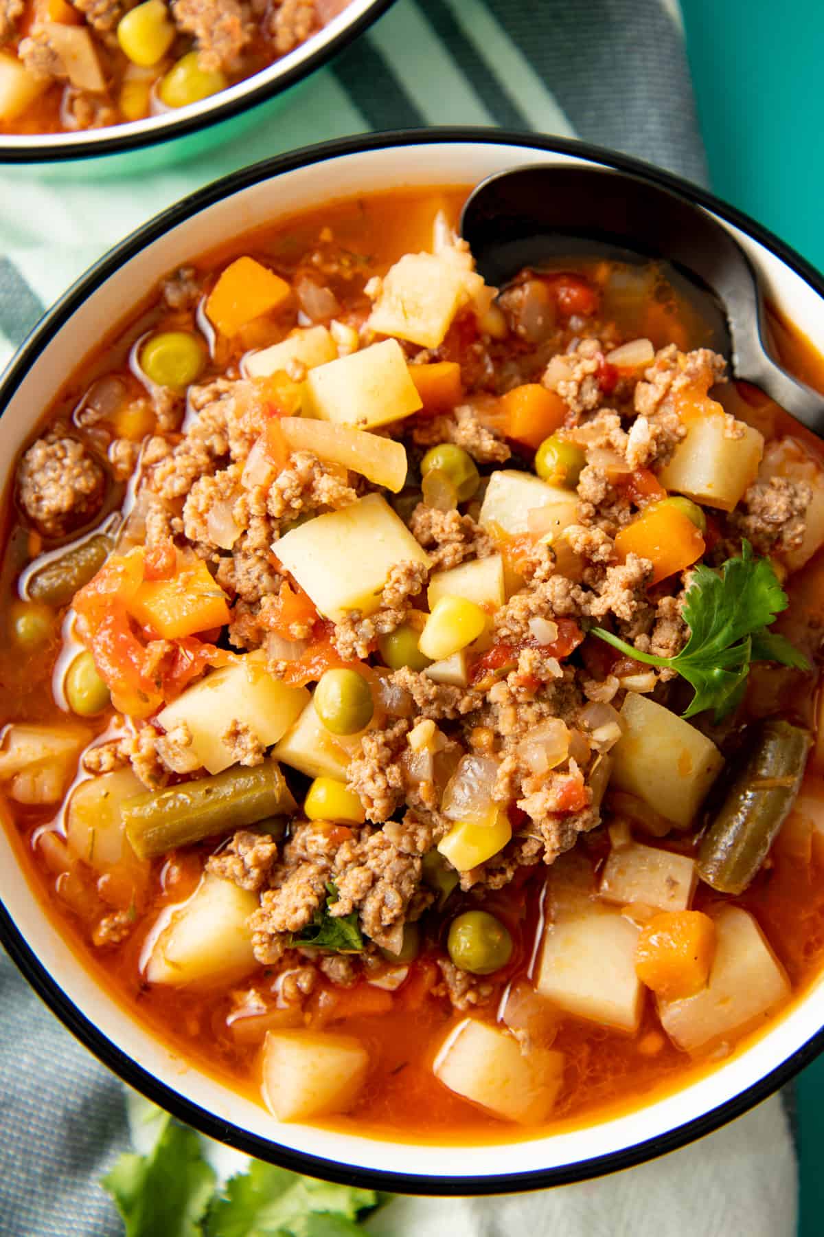 A soup filled with vegetables, potatoes, and beef fills a white bowl with a black rim.