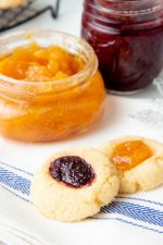 Close-up of two jam thumbprint cookies with open jars of homemade jam behind them.