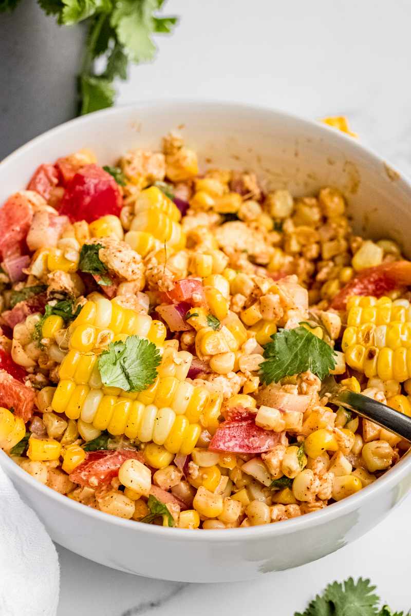 A spoon digs into a serving bowl of mexican street corn salad garnished with fresh cilantro leaves.