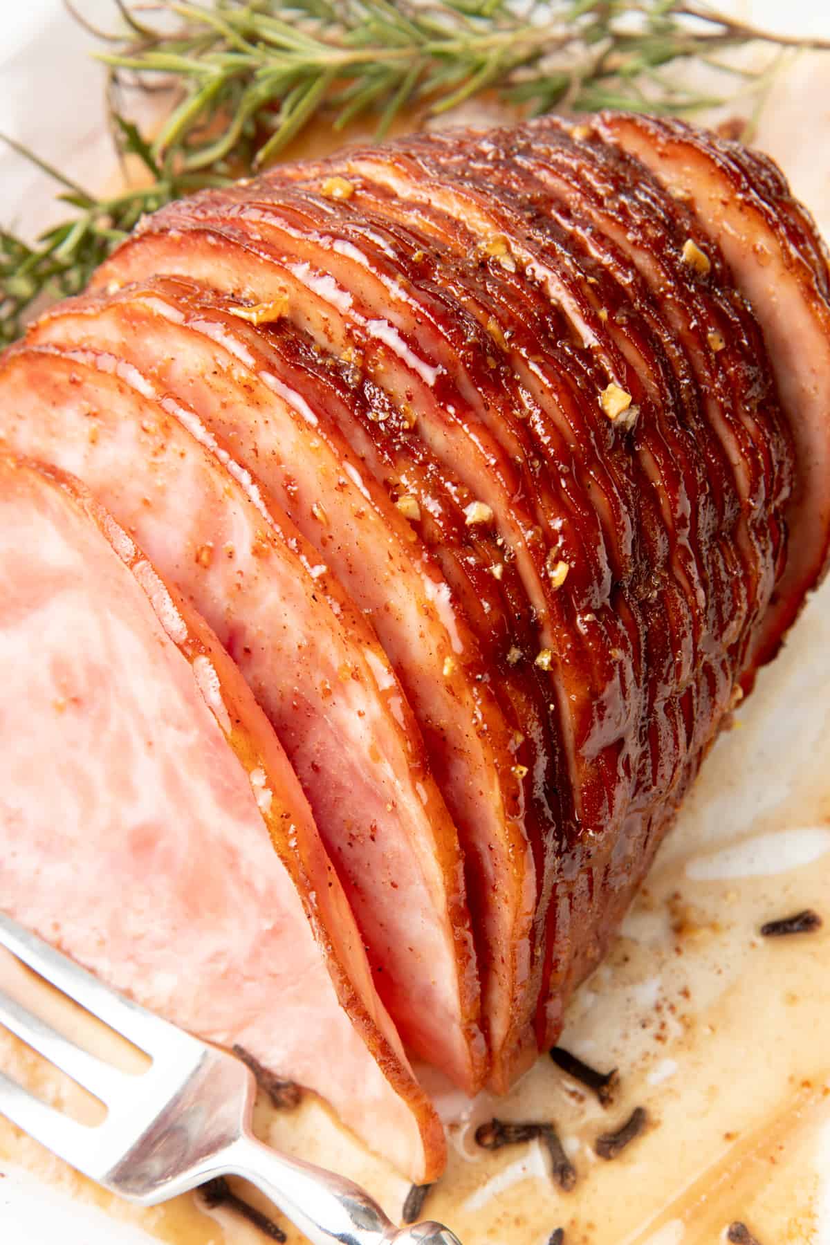 Sliced ham is glazed on a white dish. A silver fork and a sprig of rosemary also sit on the dish.