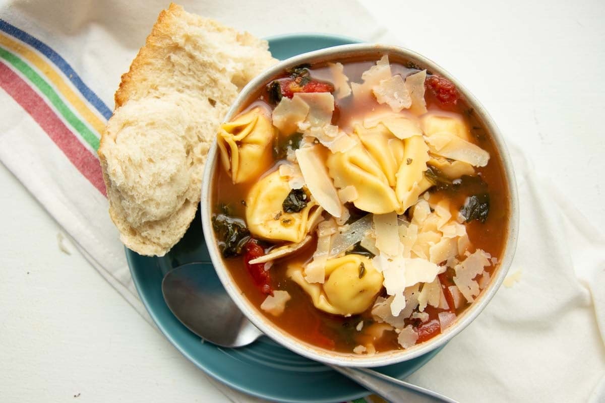 A bowl of tortellini soup garnished with shaved parmesan rests on a plate atop a striped kitchen linen.