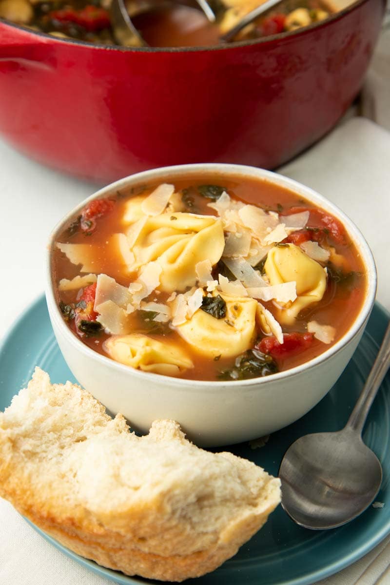 A white bowl of tortellini soup rests on a teal plate with a spoon and piece of bread alongside.