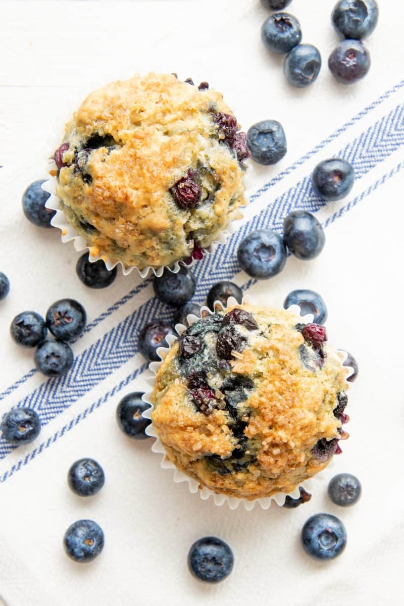 Two Vegan Blueberry Muffins sit together surrounded by fresh blueberries.