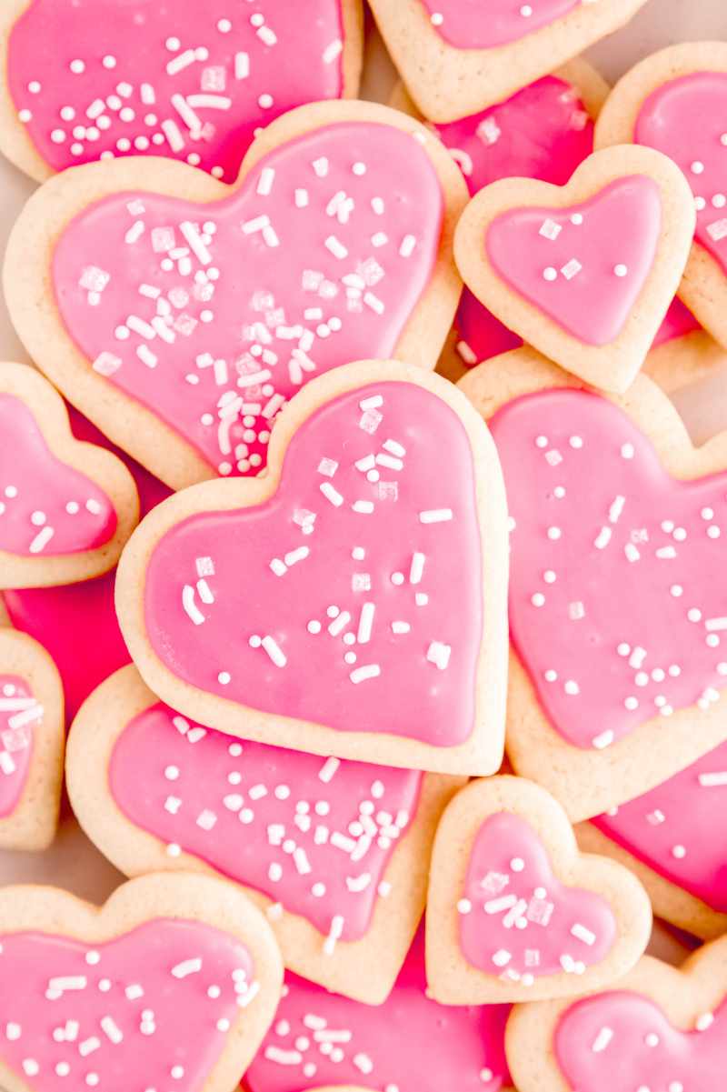 Top view of a pile of valentine's day sugar cookie hearts in two sizes topped with pink icing and white sprinkles.