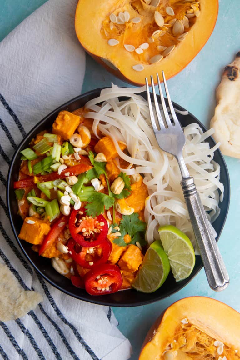 Vegan one pot pumpkin curry served over a bed of rice noodles, in a black dish on a light blue table.